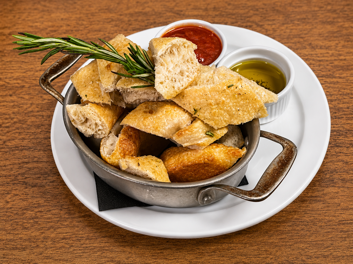 Bowl of cooked bread pieces garnished with rosemary, with side of marinara sauce and olive oil on a white plate.