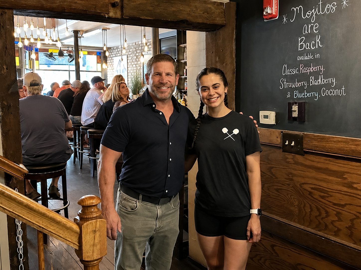 Two people, a man and a woman, standing together inside a bar or restaurant, smiling at the camera. The man has short hair, a beard, and is wearing a dark button-up shirt and light-colored pants. The woman has long dark hair styled in a braid, wearing a black t-shirt with two crossed sticks on it and black shorts. There are other patrons seated at the bar behind them, and a chalkboard on the wall to the right advertising the return of mojitos in various flavors.