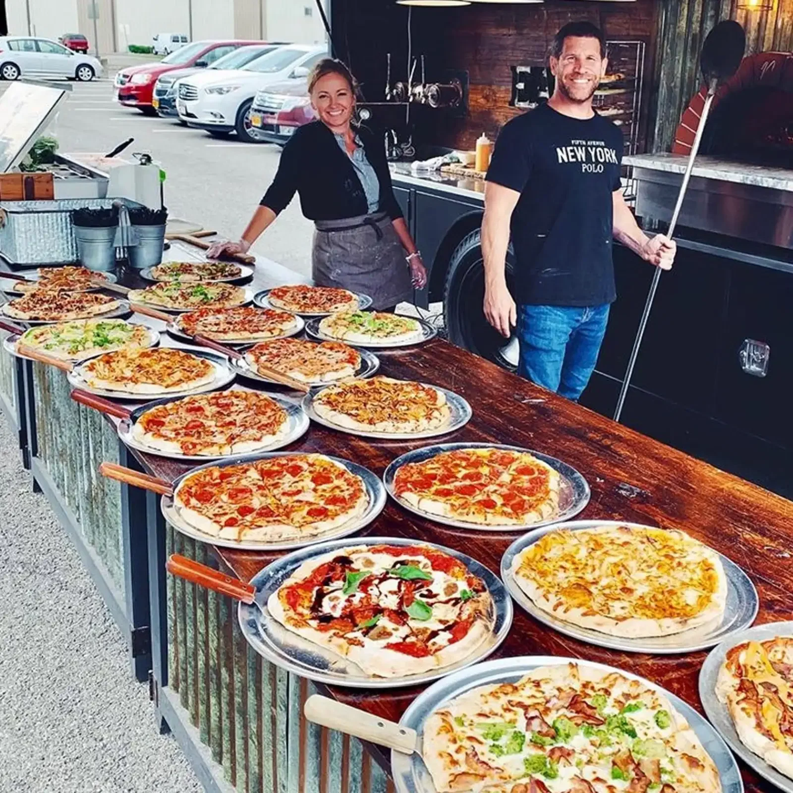 Outdoor pizza stand with numerous pizzas on platters, two smiling vendors, parked cars in the background.