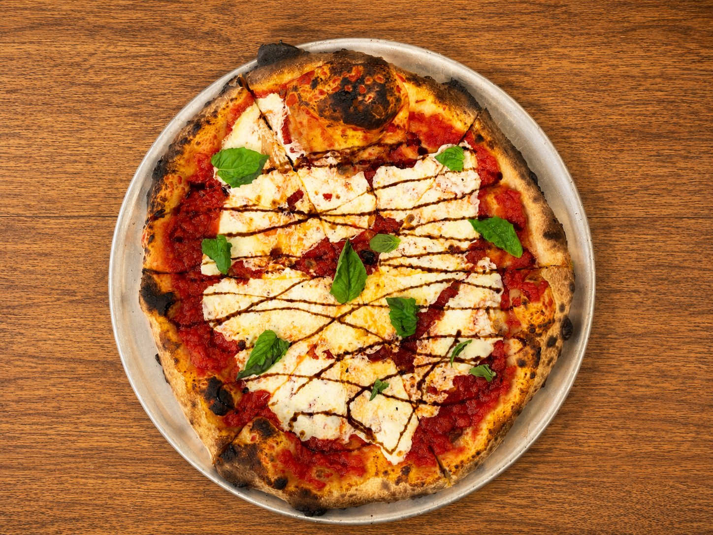 A cheese pizza with basil leaves, tomato sauce, shredded cheese, and drizzle of balsamic glaze on a round metal tray on a wooden table.