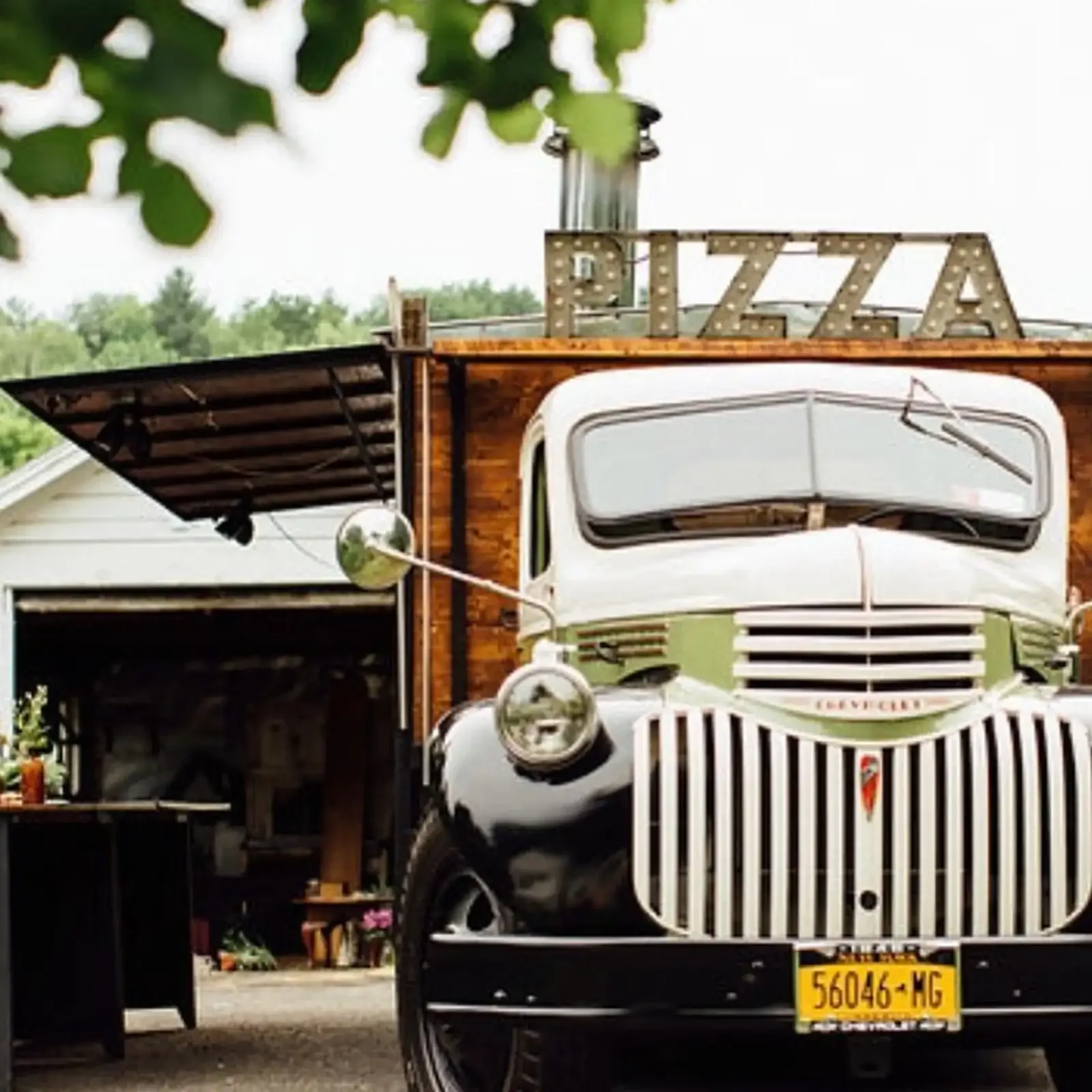 A vintage white truck with green accents and a yellow license plate standing in a yard with a wooden structure and a sign that reads 'PIZZA' lit with marquee lights on top. Green trees are in the background.