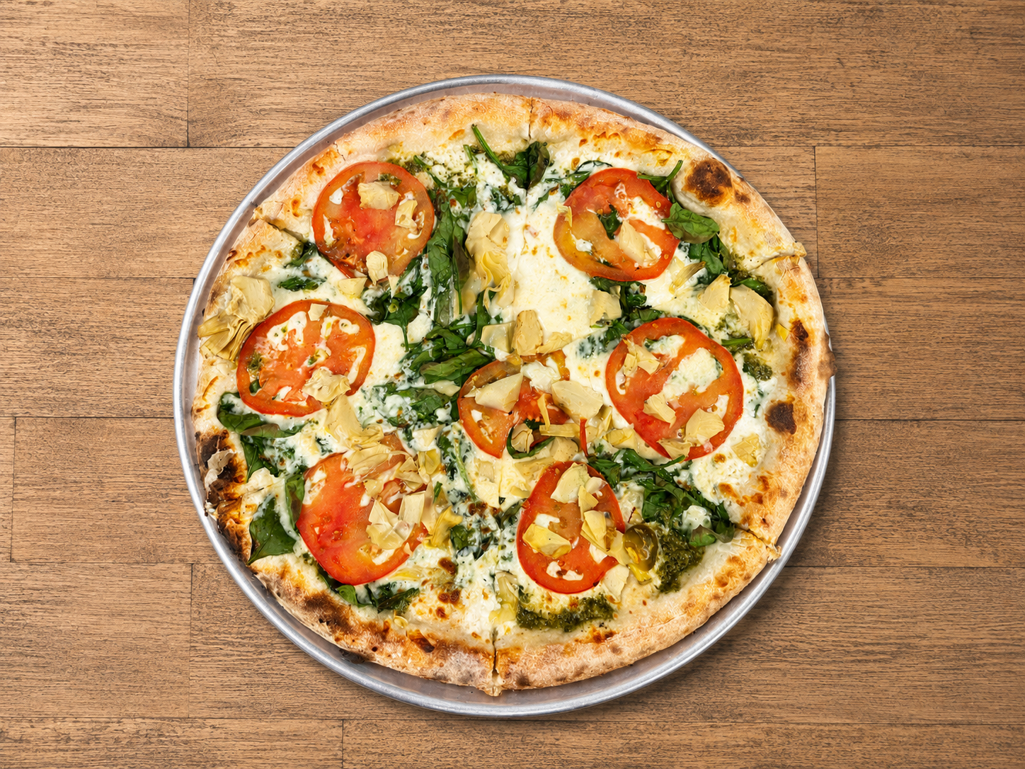 A cheese pizza topped with tomato slices, basil, artichoke hearts, and green sauce on a silver tray, on a wooden table.