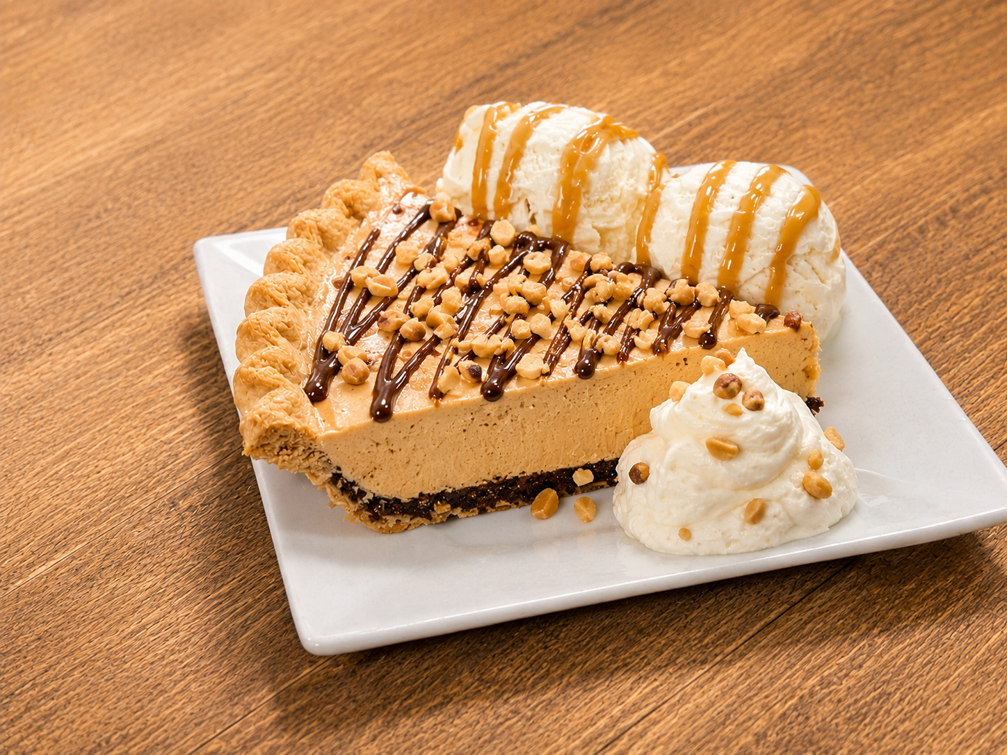 Slice of peanut butter pie with chocolate drizzle and chopped peanuts, served with two scoops of vanilla ice cream topped with caramel sauce and whipped cream with peanuts, on a white square plate on a wooden table.
