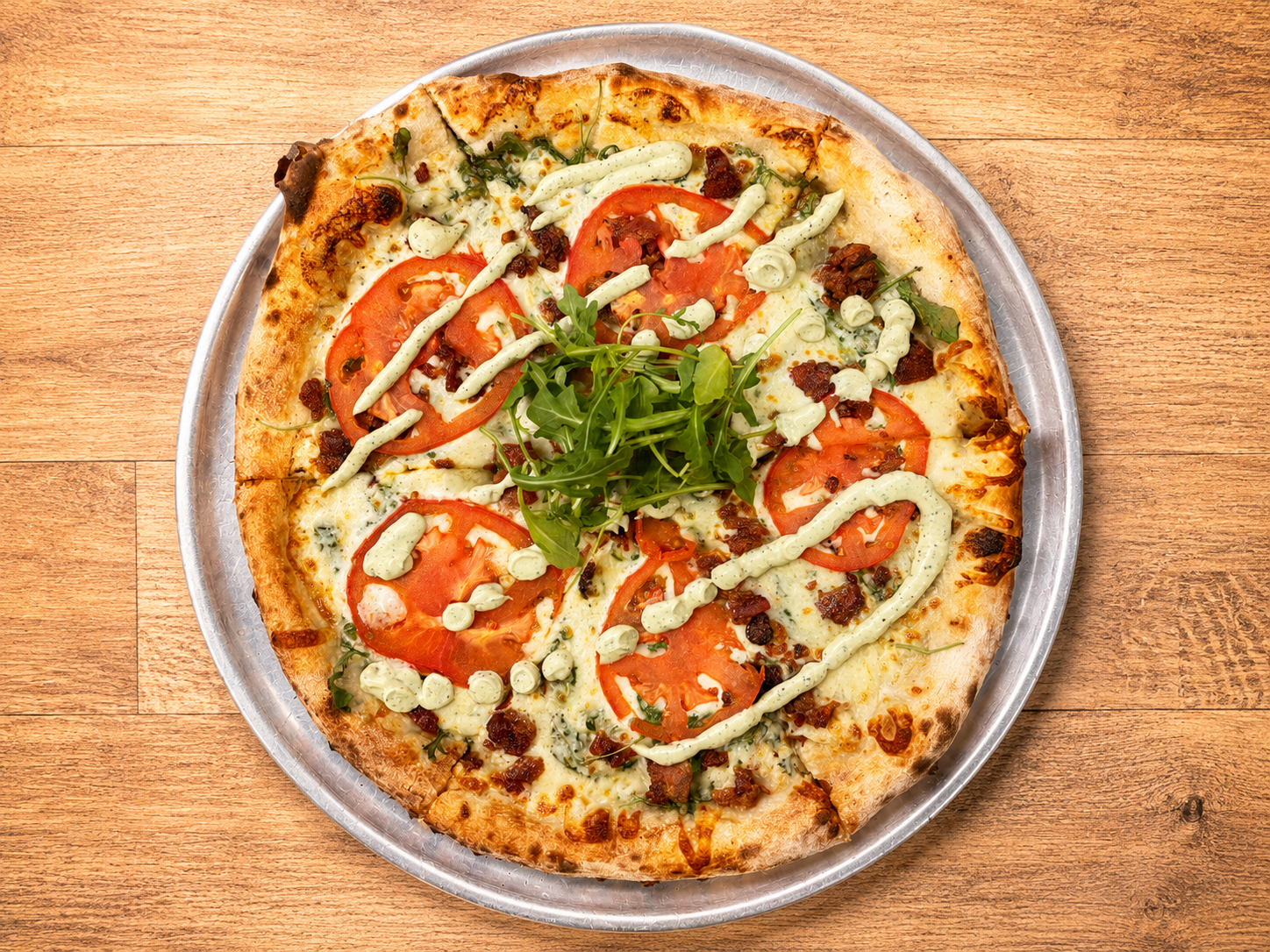 Pizza topped with sliced tomatoes, arugula, green sauce, and cheese on a wooden table.