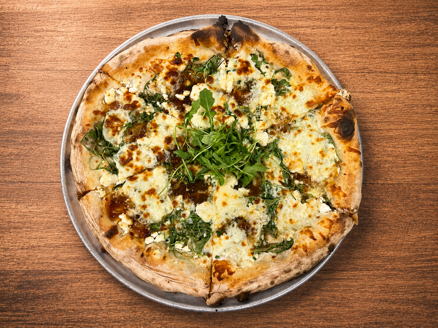 A pizza with cheese, arugula, and balsamic glaze on a metal tray, placed on a wooden table.