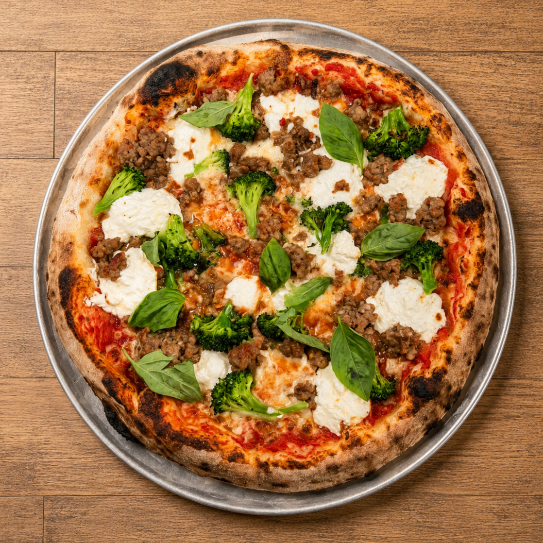 Close-up of a pizza with cheese, sausage, broccoli, and basil on a metal tray on a wooden surface.