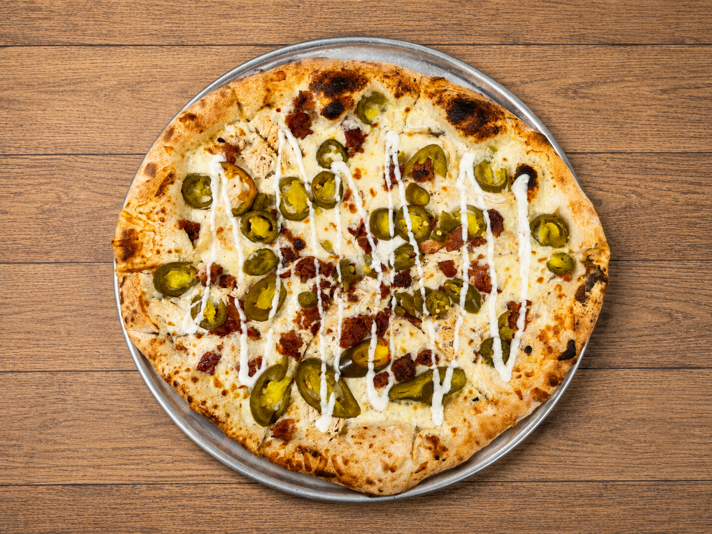 A pizza with cheese, sliced jalapeño peppers, and drizzled white sauce on a metal tray on a wooden surface.
