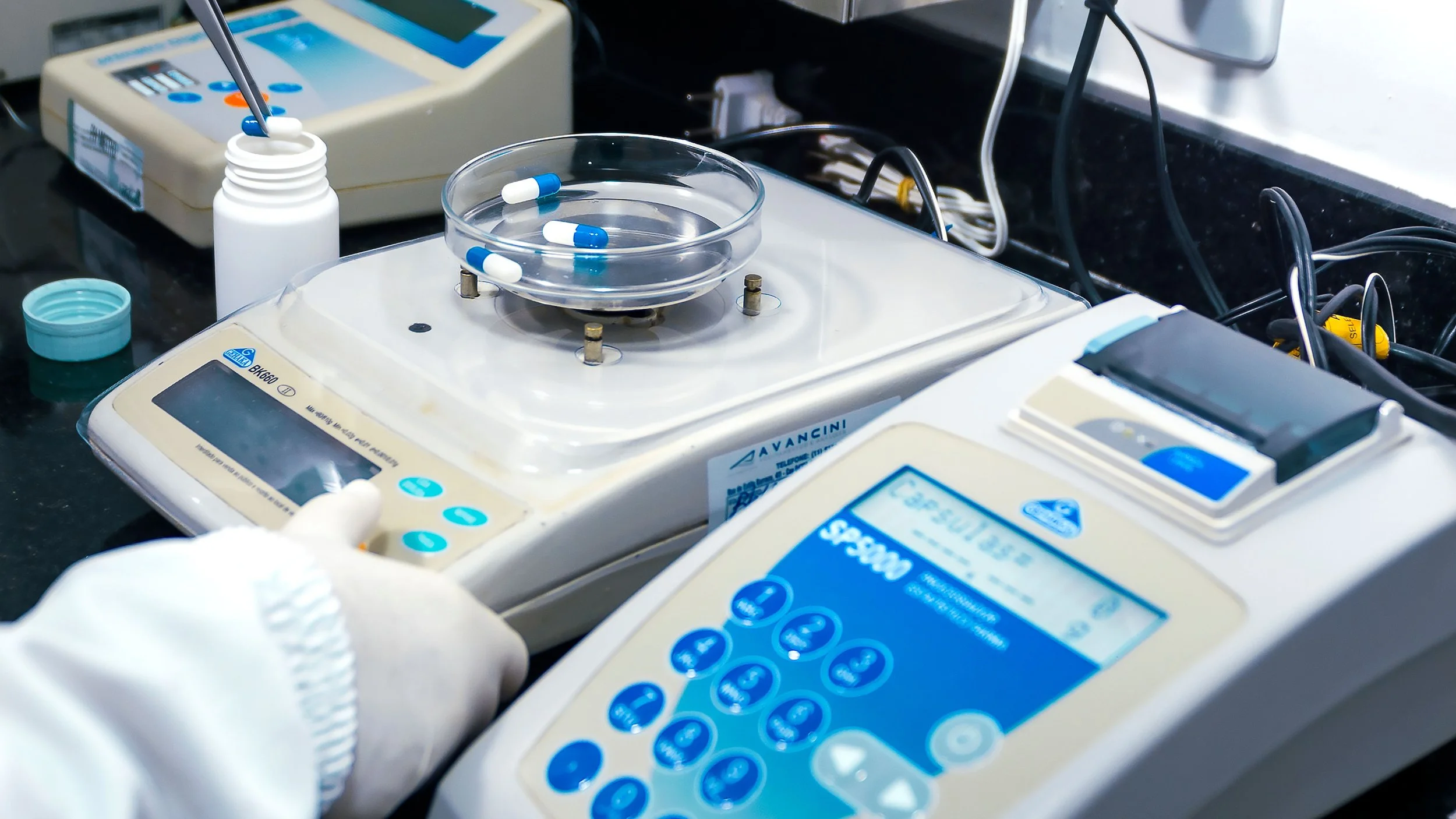 Close-up of laboratory equipment, including a centrifuge with capsules inside, a bottle of capsules, and a digital device with a blue display, set on a black work surface.