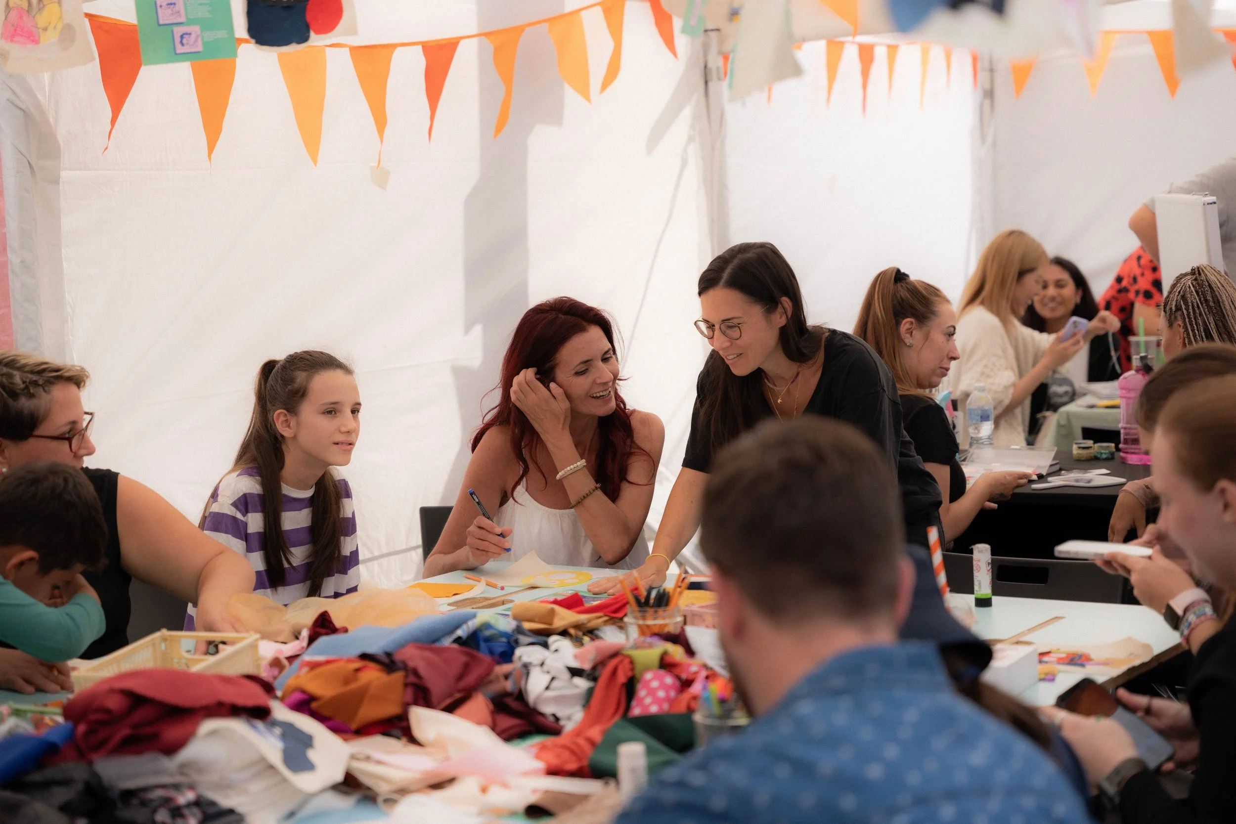 A group of children and women sitting at tables participating in a craft activity, with various craft supplies and fabric pieces on the table. Banner decorations are hanging above them.