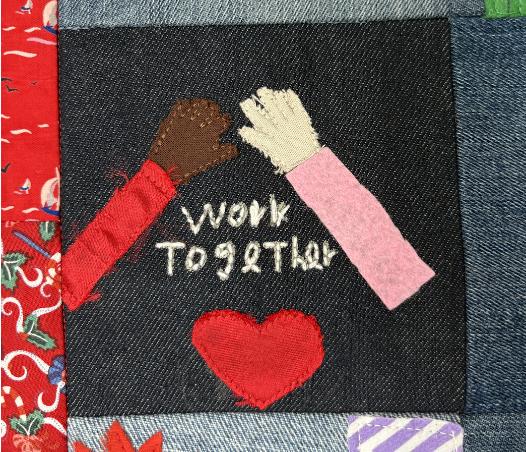 Embroidery patch with two raised hands, one brown and one white, giving a high five. Below the hands, the embroidered words "work together" are written, with a large red heart underneath. The patch is sewn onto denim fabric, surrounded by a colorful patterned fabric.