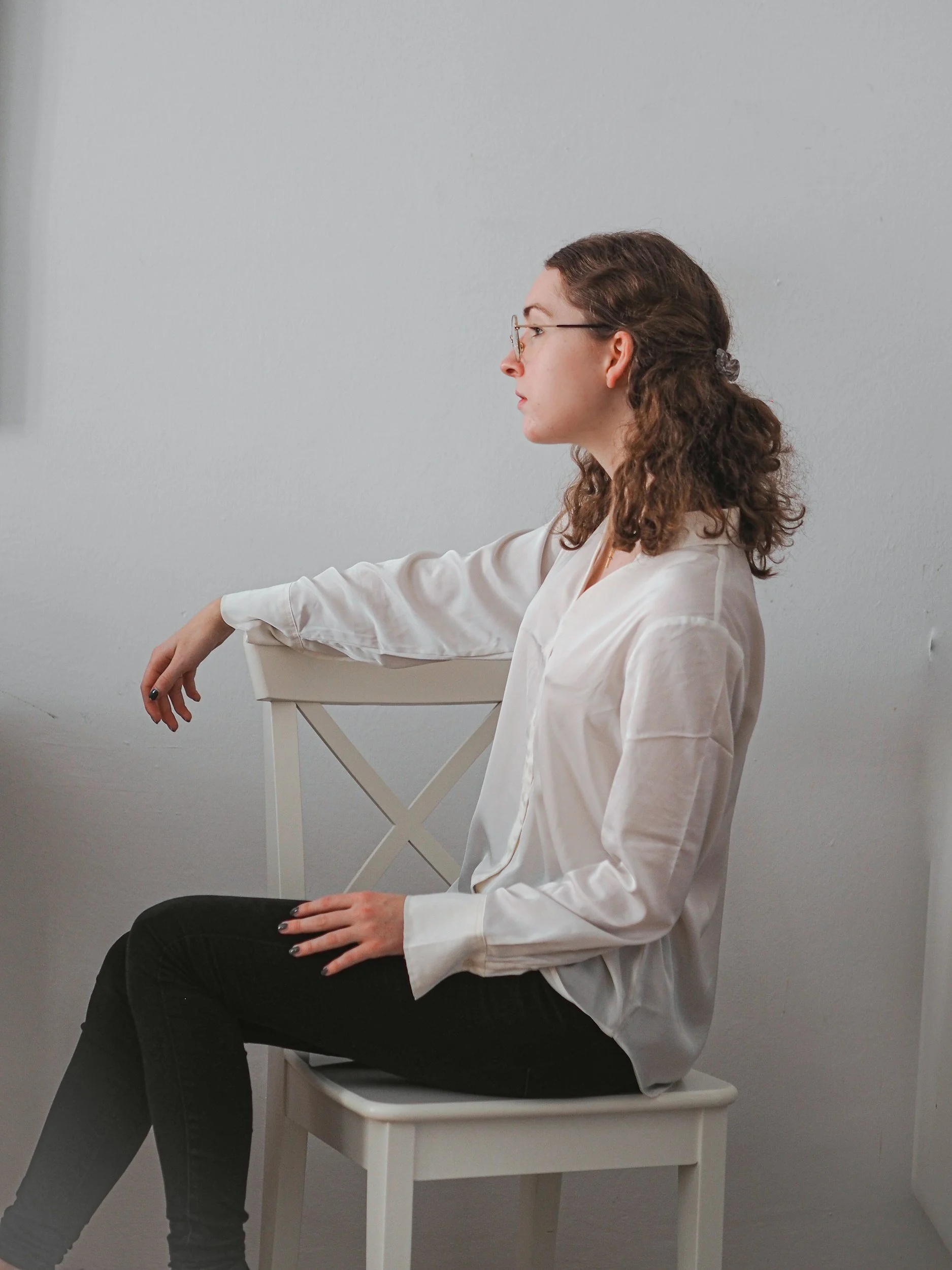 A young woman with curly brown hair, wearing glasses and a white blouse, sitting on a white chair against a plain light gray wall, with her head turned to the side.