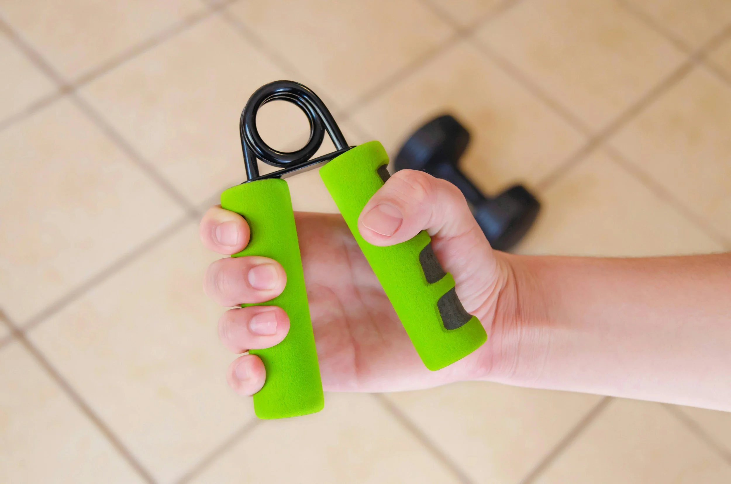 A person holding a green-handled grip strengthener with black coiled springs, standing on a beige tiled floor.