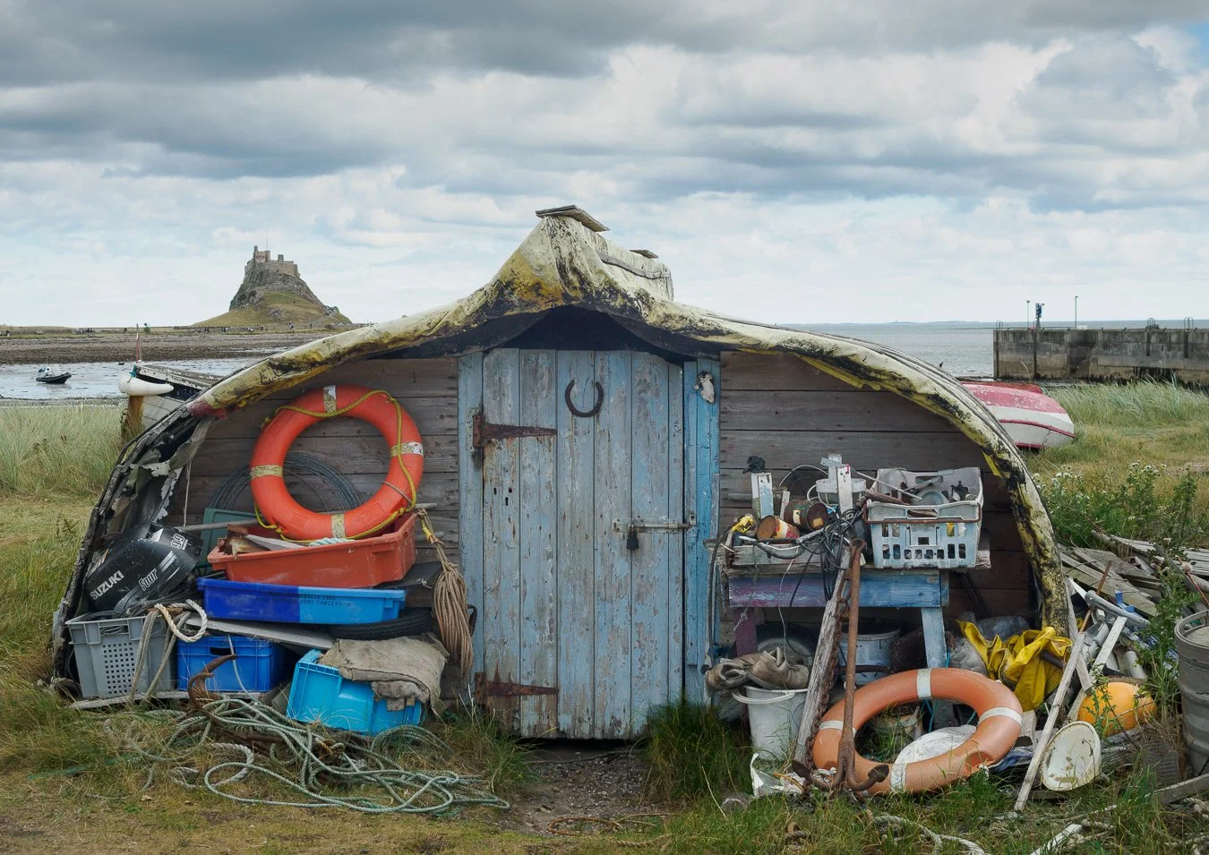 Small weathered boat shed filled with boating gear, life rings, ropes, and tools, set on grassy ground near a harbor with boats and a castle on a hill in the background under cloudy sky.