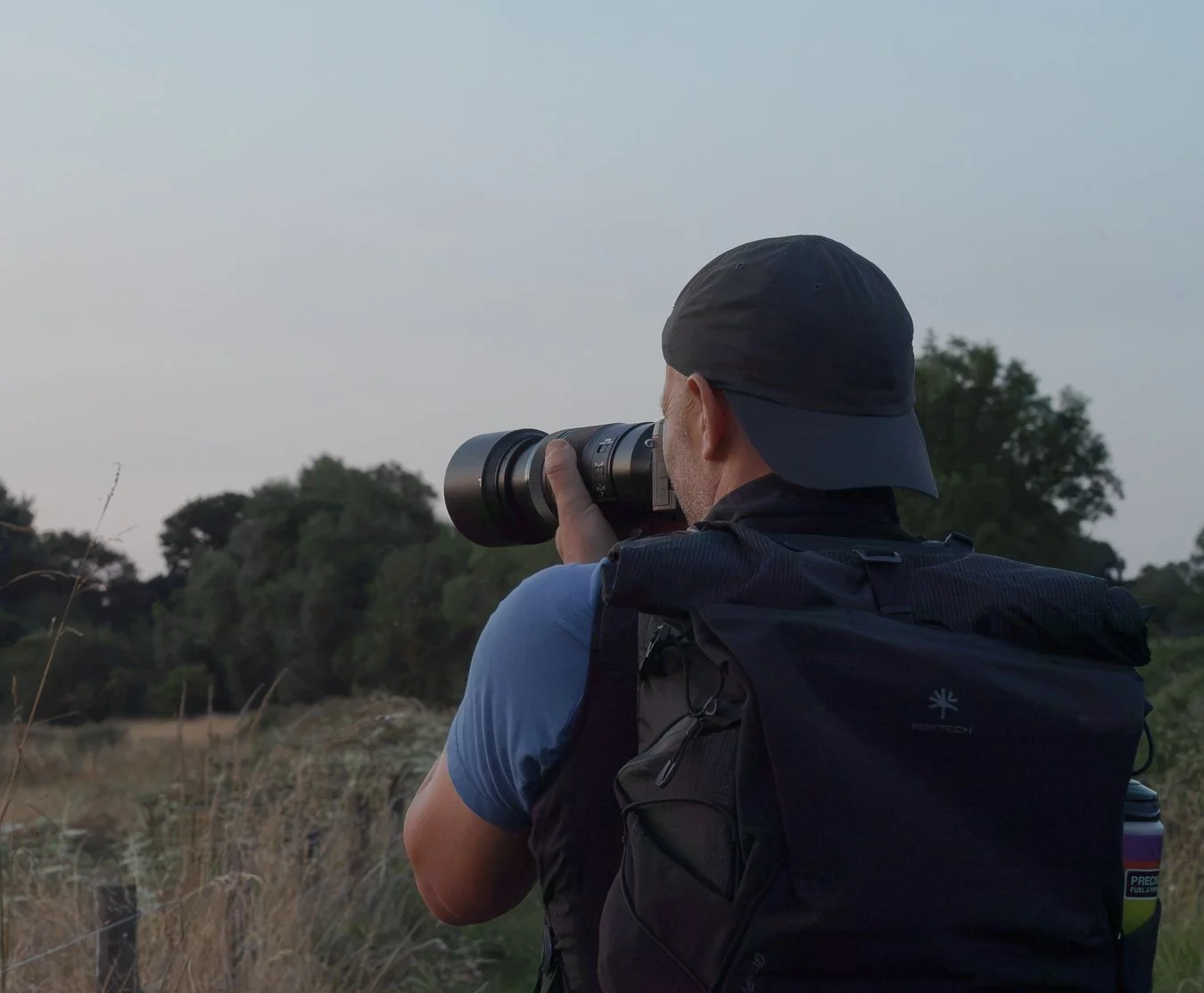 A person wearing a black cap and blue shirt is taking a photograph with a telephoto lens in an outdoor, rural area with trees and grass.