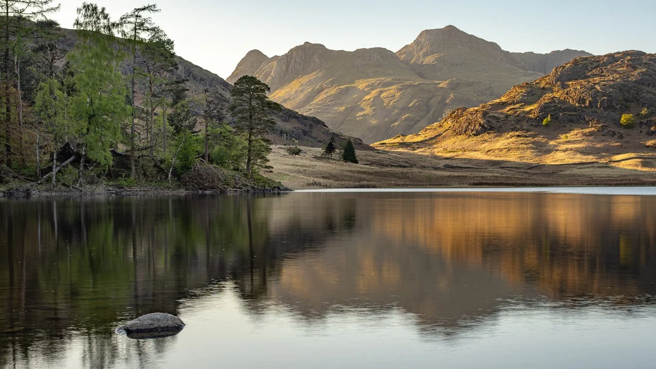 Peaceful mountain landscape with a calm lake reflecting the surrounding hills and trees, illuminated by soft sunlight.