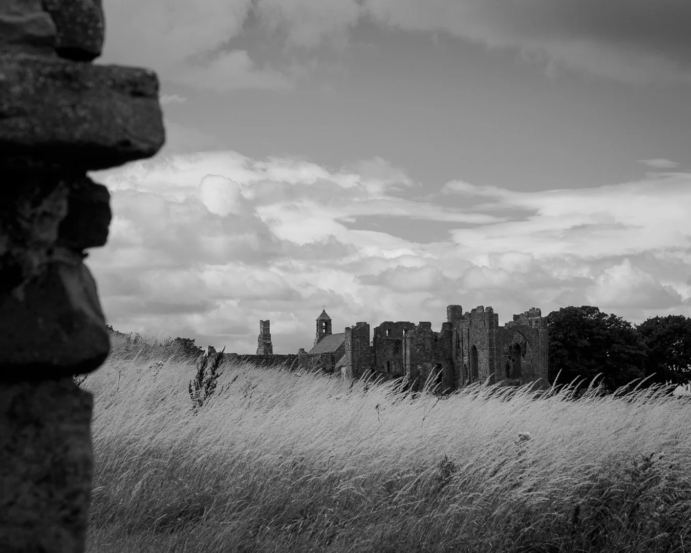 Black-and-white photo of an abandoned castle in a grassy field, with a stone wall in the foreground and a cloudy sky above.