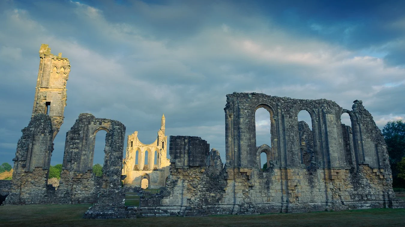 Ruins of an old stone building with large arched windows, set against a partly cloudy sky with scattered clouds and a grassy foreground.