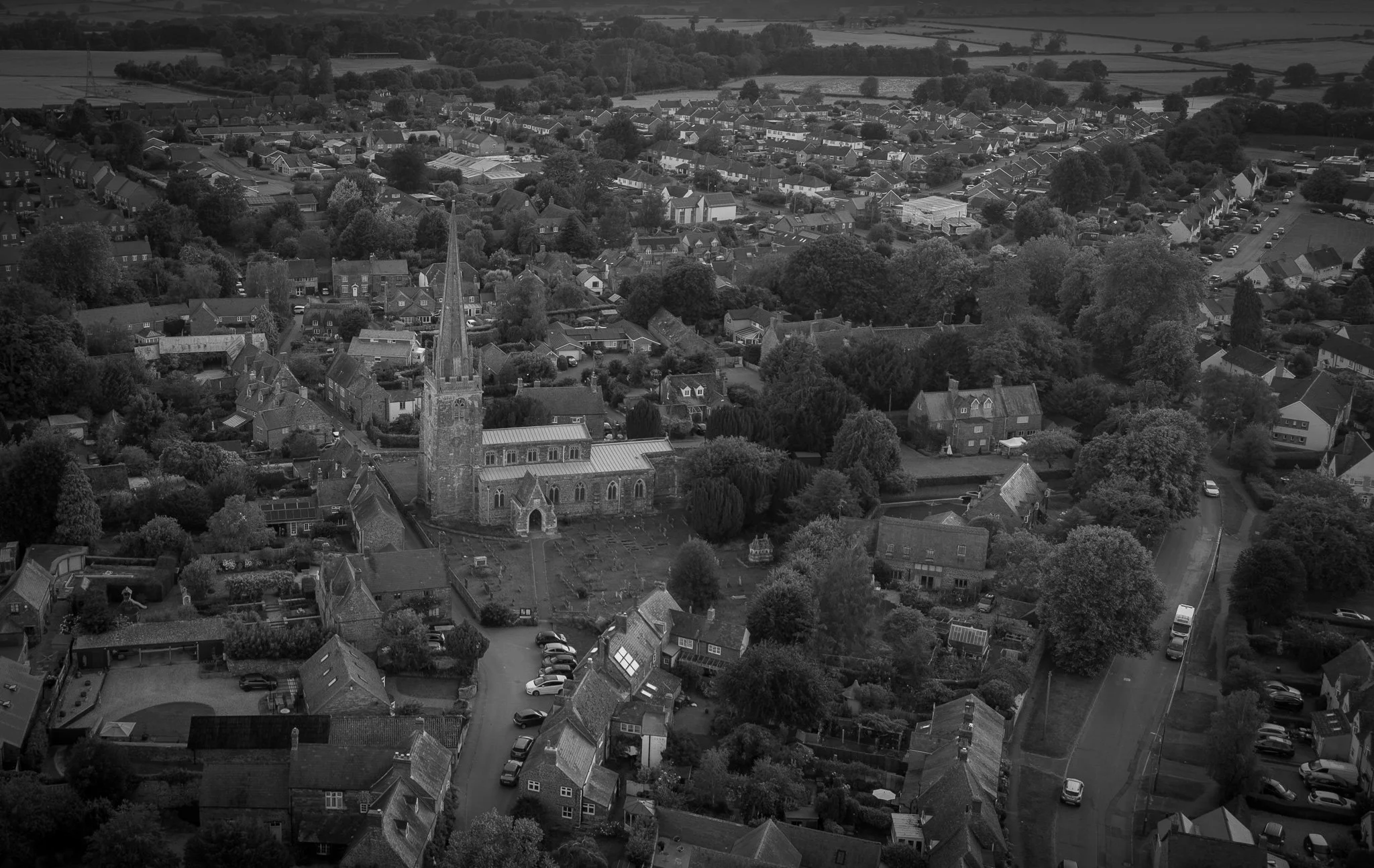 A grayscale aerial view of a small town with a church featuring a tall steeple in the foreground, surrounded by houses, trees, roads, and open fields in the background.