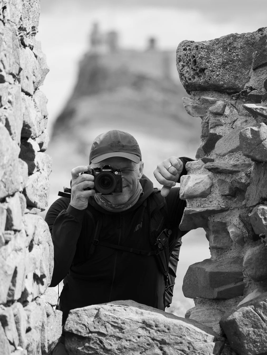 A man taking a photo with a camera through a stone window or opening in a wall, with a building or castle in the background.
