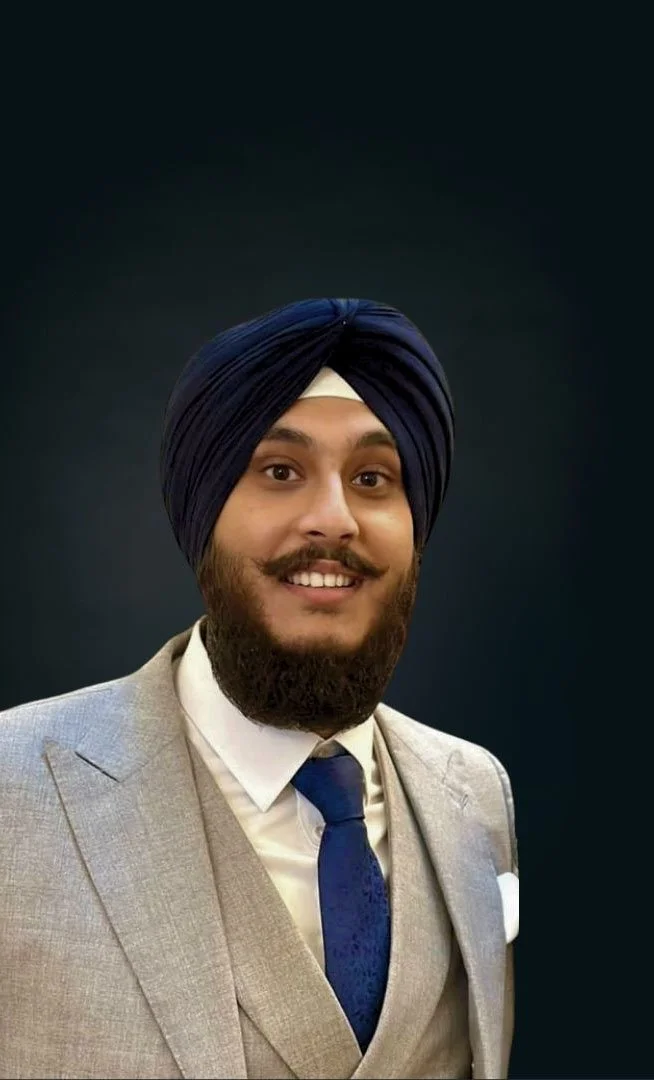 Portrait of a man wearing a navy blue turban, a light gray suit, white shirt, and navy blue tie, smiling against a dark background.