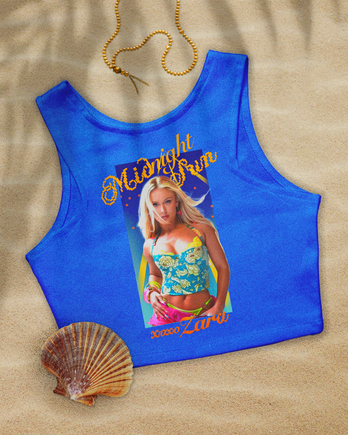 A blue tank top with a graphic of a woman in a floral top and pink shorts, with the text 'Midnight Zone' and 'Xoxo Zara' on it. The tank top is on a sandy surface, alongside a seashell and a gold beaded necklace.