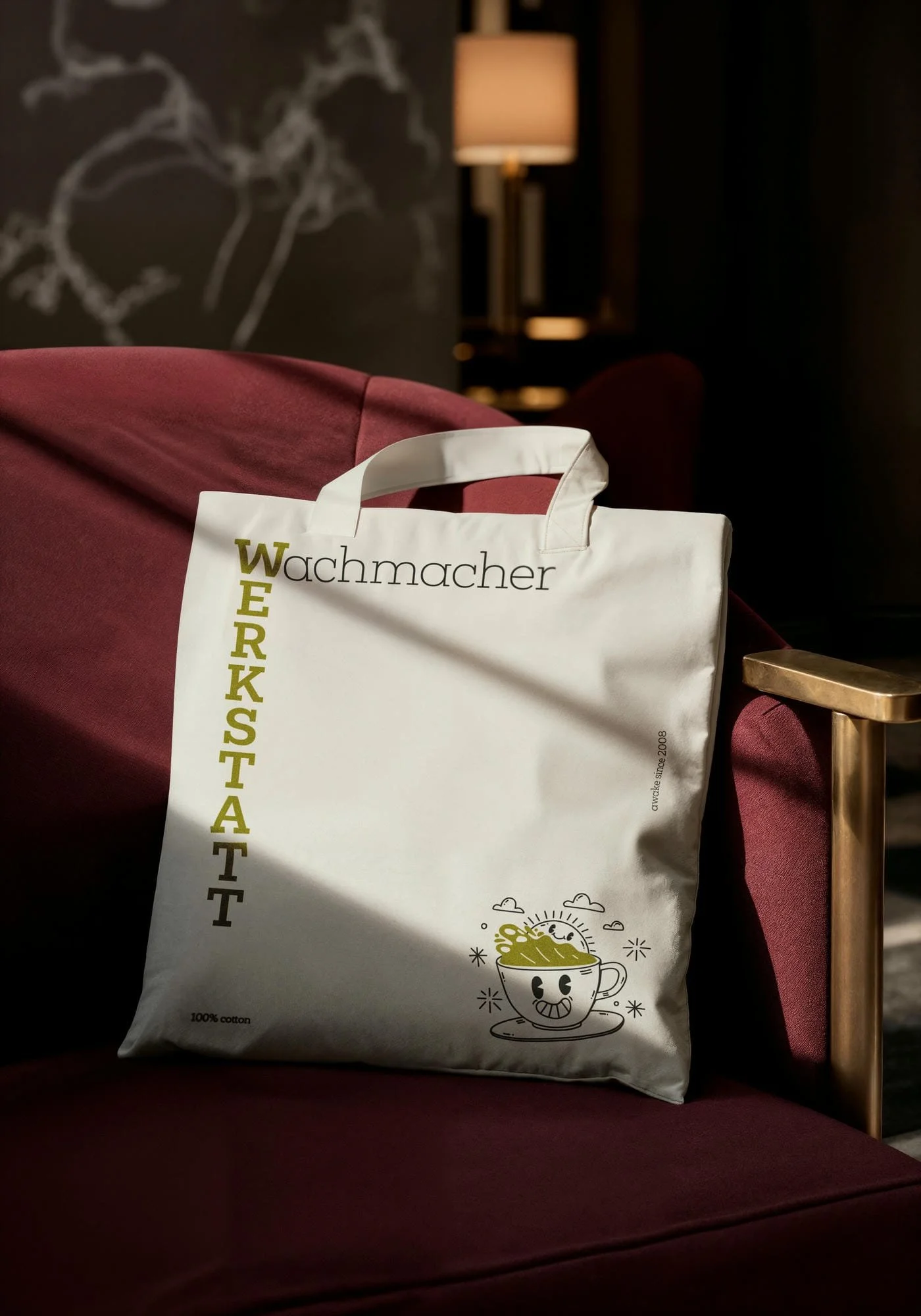 Eine weiße Einkaufstüte mit einer Zeitung, die auf einem dunkelroten Sessel liegt. Im Hintergrund ist eine Stehlampe und eine Wand mit einer schwarzen Tafel und weißen Zeichnungen. Die Tüte hat ein lustiges Kaffeebild und die Wörter »Wachmacher« und 