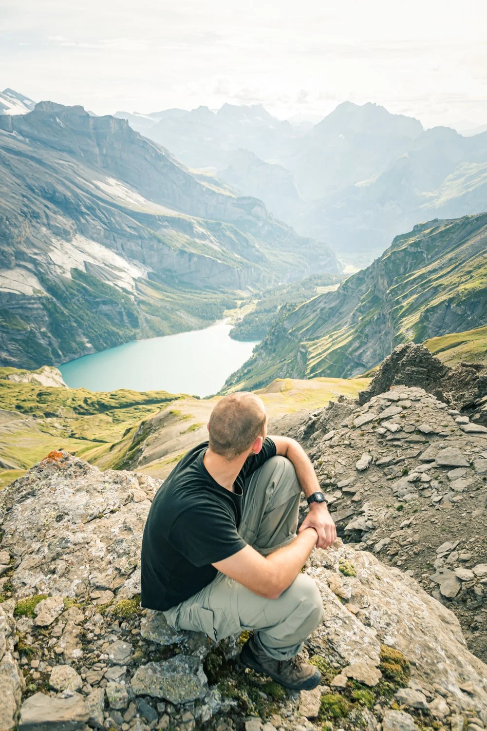 Ein Mann sitzt auf einem Felsen und blickt auf eine Berglandschaft mit Wasser und Tälern.