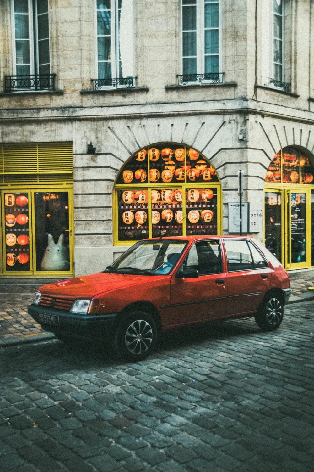 Ein rotes Auto parkt auf einer gepflasterten Straße vor einem Gebäude mit gelben Fensterrahmen und Beleuchtung, die mit japanischen Laternen geschmückt sind.