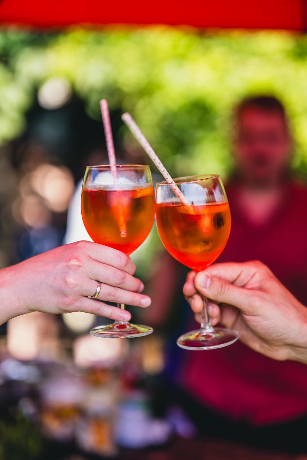 Zwei Menschen stoßen mit Gläsern auf ein Fest an, die Gläser enthalten ein rotes Getränk mit Strohhalmen. Es wird im Freien gefeiert, im Hintergrund sind unscharfe Personen und grüne Bäume zu sehen.