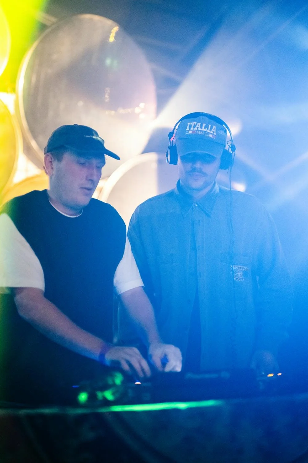 Zwei Männer arbeiten an einem DJ-Setup, einer trägt eine Mütze mit italienischer Flagge und das Wort 'ITALIA', der andere einen schwarzen Cap, beide mit Kopfhörern, im Hintergrund große silberne Hörner und bunte Lichteffekte.