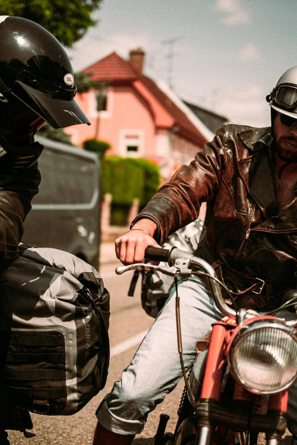 Zwei Männer auf Motorrädern im Gespräch, einer trägt einen schwarzen Helm und der andere eine Lederjacke mit Schutzbrille, im Hintergrund Häuser und Bäume.