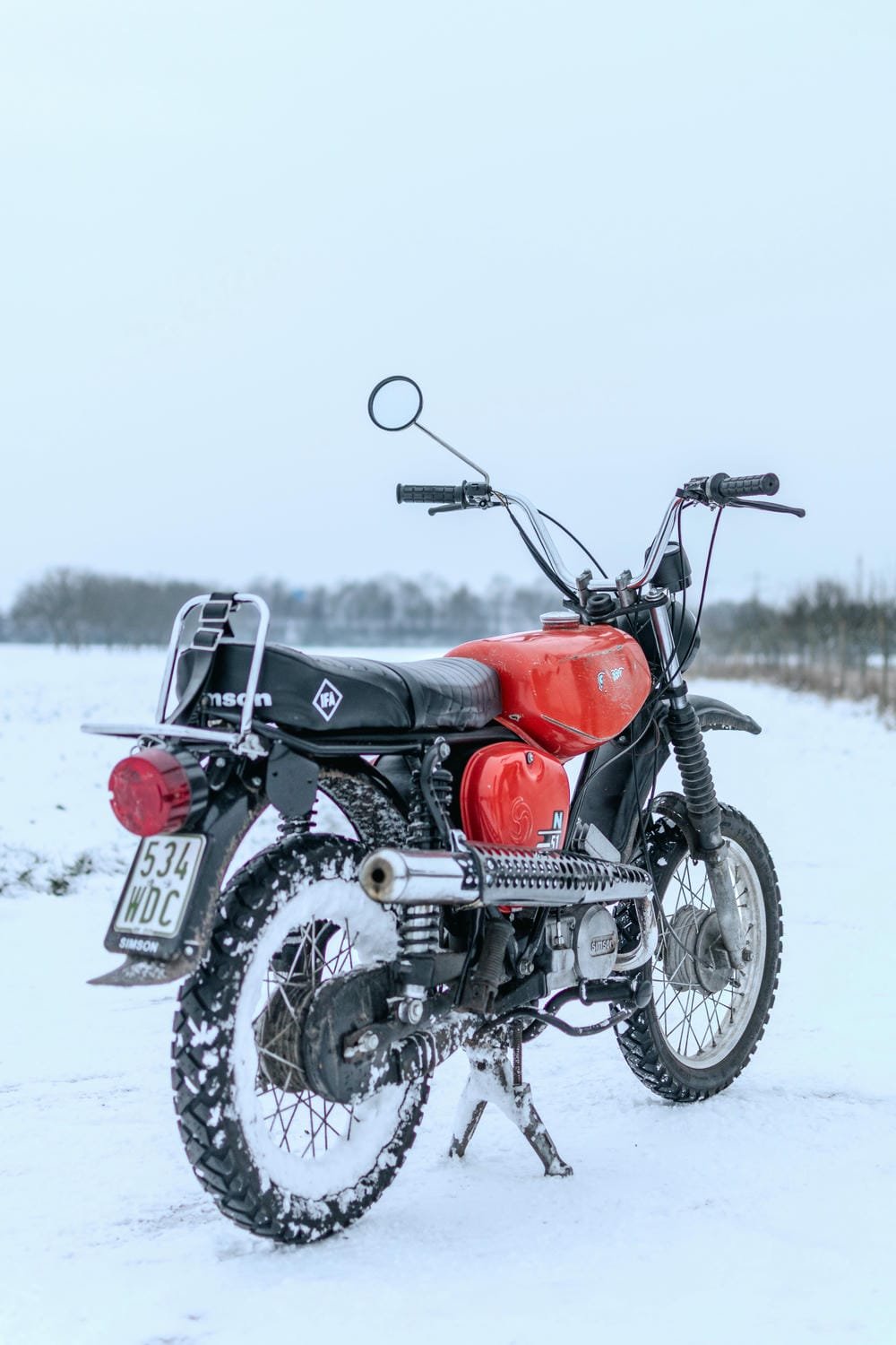 Rote Vintage-Motorrad steht auf verschneitem Feld, der Himmel ist grau und wolkenverhangen.
