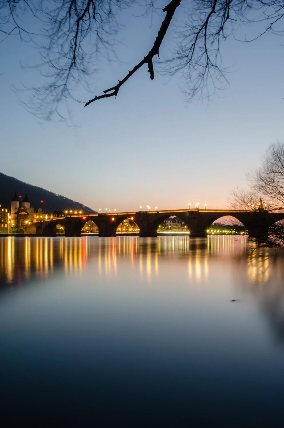 Blick auf eine beleuchtete Brücke über einen Fluss bei Dämmerung, mit reflektierenden Lichtern im Wasser und Baumzweigen im Vordergrund.
