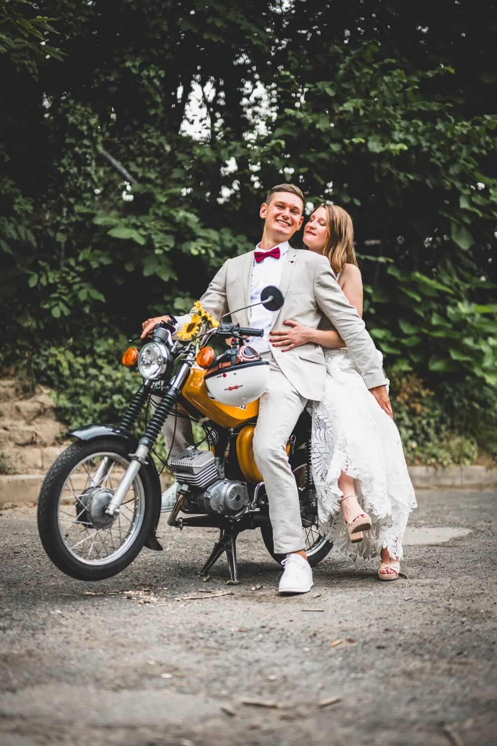 Ein lachendes Paar in formeller Kleidung sitzt auf einem gelben Motorrad vor grünen Bäumen.