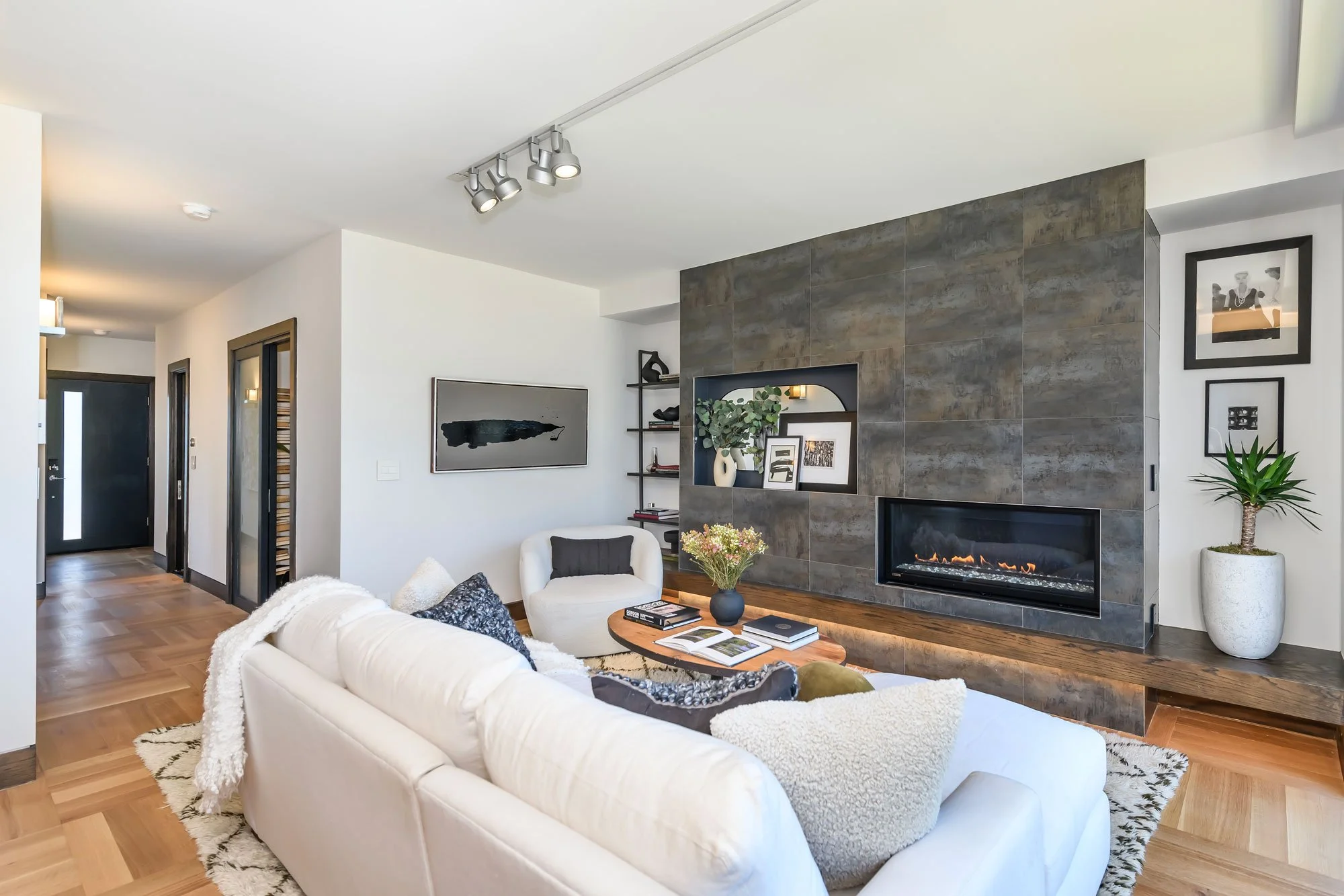 Modern living room with white sofa, black and white decorative pillows, wooden coffee table with open books, a fireplace on a tiled wall, green potted plant, framed artwork, and professional track lighting on ceiling.