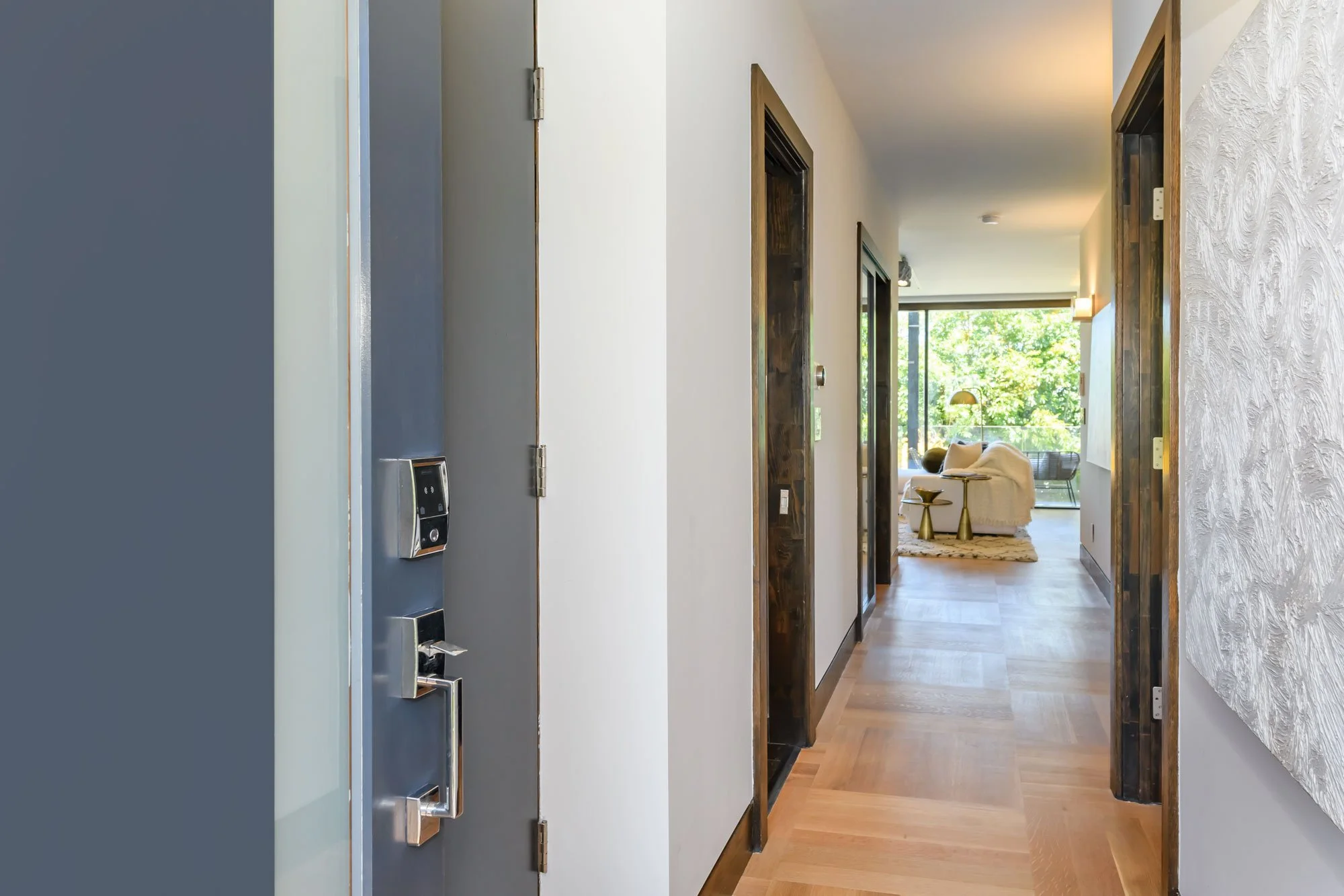 View of a modern apartment hallway leading into a living room with large windows, a white couch, and gold side tables, with wooden floor and trim.