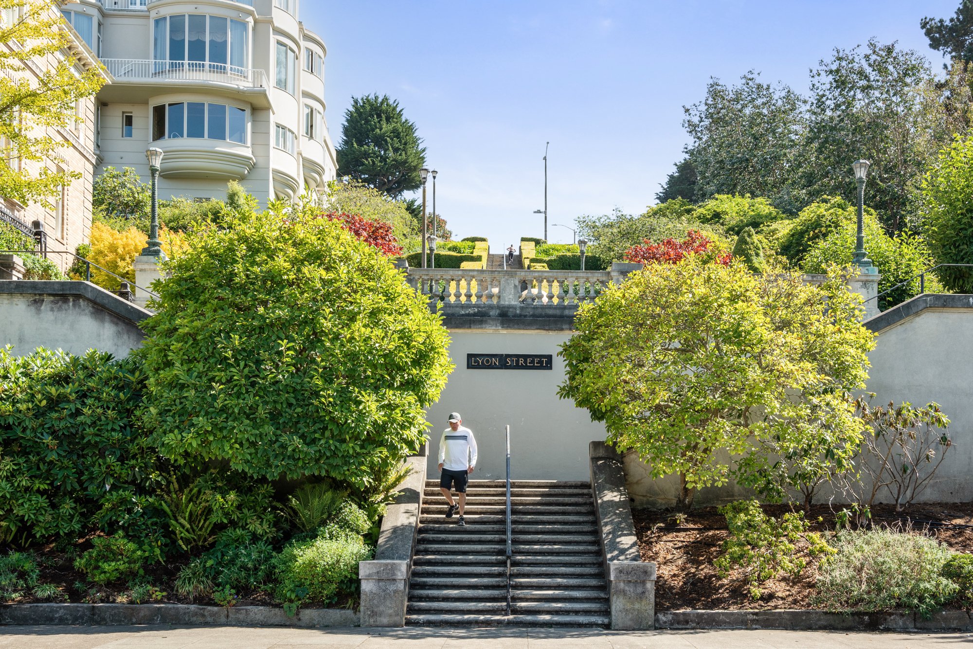 A man walking down the stairs on Lyon Street near lush green trees and bushes; a building with rounded windows can be seen on the left, and there are steps leading up to a walkway with lamp posts and a person standing at the top.