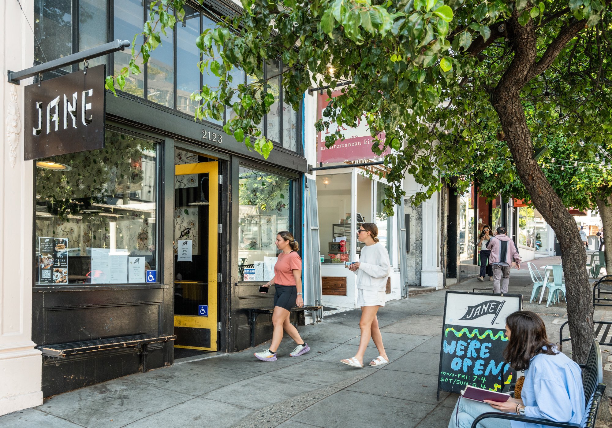 Street scene outside a restaurant named JANE with people walking and sitting at outdoor tables under trees. A woman in white is walking past the restaurant, and others are sitting nearby, some looking at their phones. A chalkboard sign indicates the 