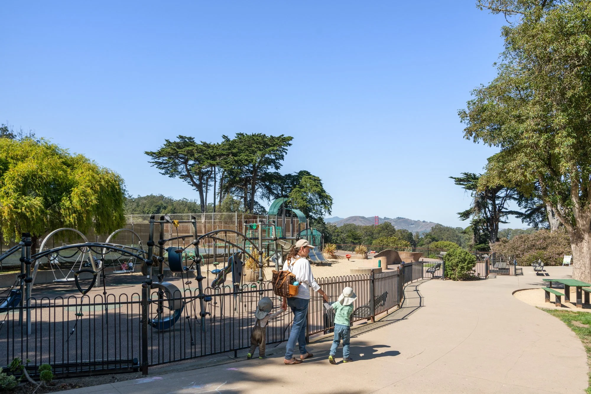 A sunny day at a playground with trees surrounding it. Children and a woman with a hat and backpack are walking beside a fenced area with playground equipment and slides, with the Golden Gate Bridge visible in the background.