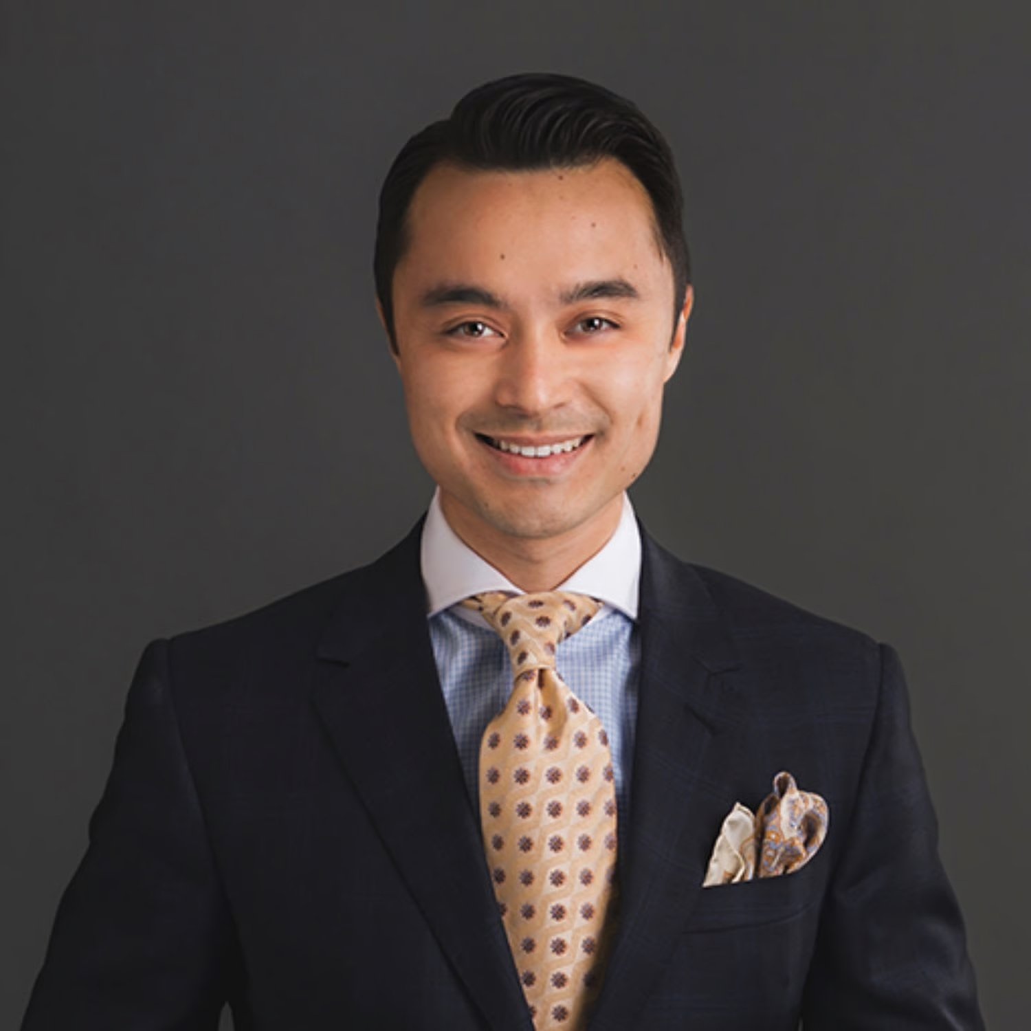 Portrait of a smiling man in a business suit with a dress shirt, patterned tie, and pocket square, standing against a dark background.