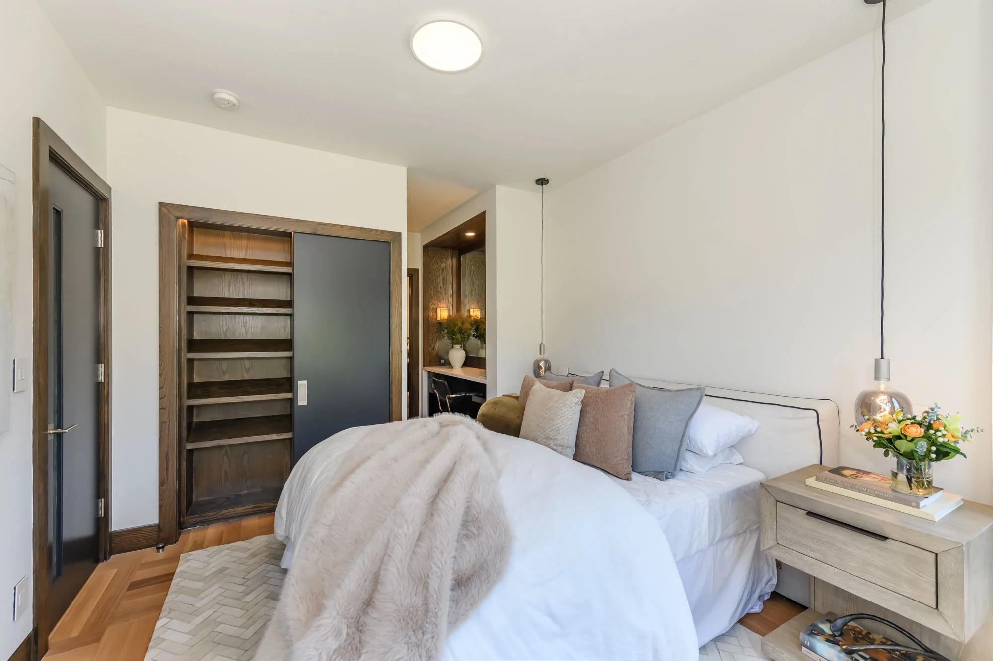 A cozy bedroom with a large bed covered in white bedding and pillows, a twin nightstand with books and a flower vase, a cozy seating area with a sofa, bookshelf, and modern pendant lighting, and minimal decor with a plant and soft lighting.