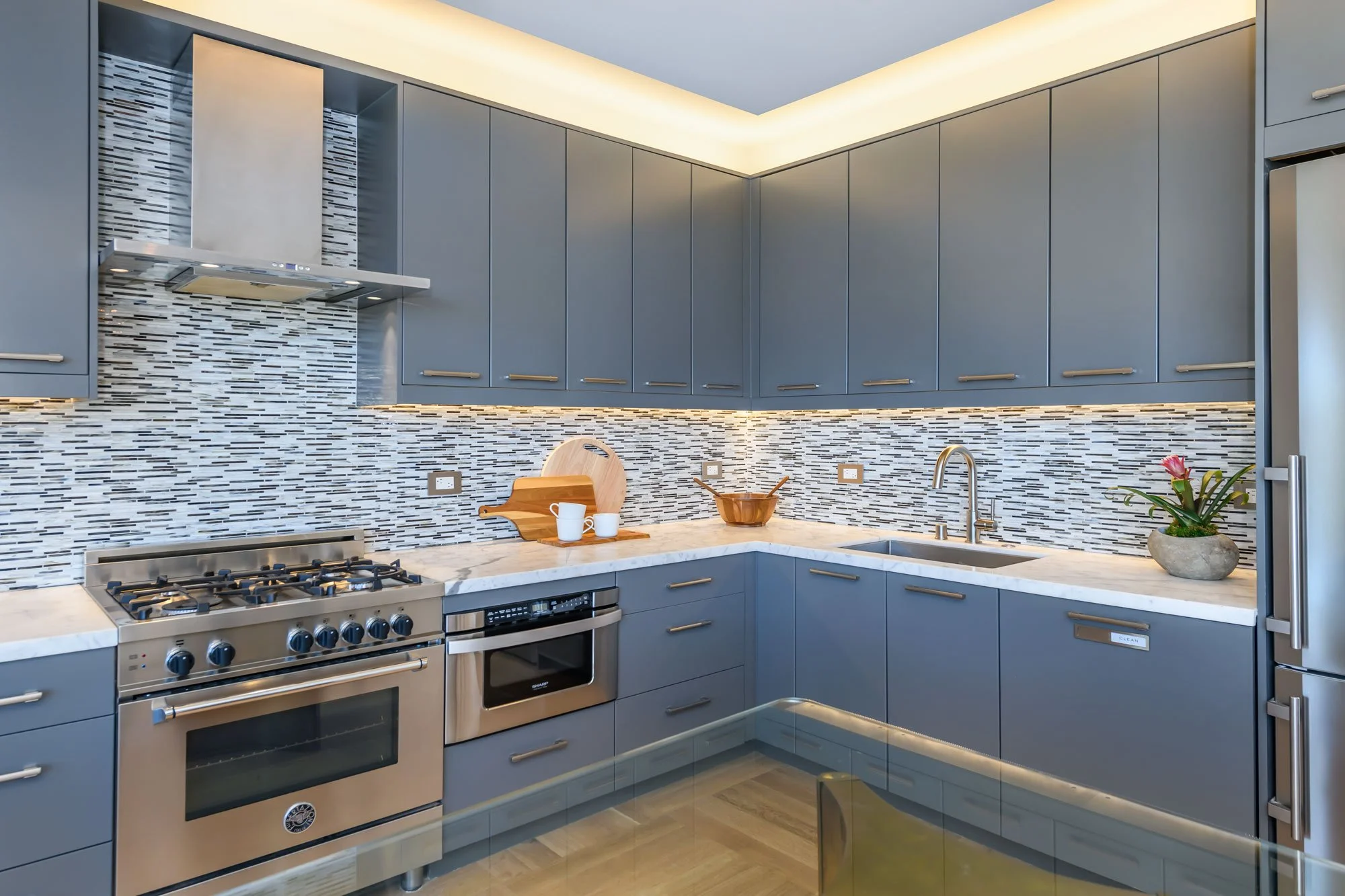 Modern kitchen with gray cabinets, a marble countertop, a stainless steel stove, oven, and range hood, a decorative mosaic tile backsplash, a small potted plant, and a corner sink.