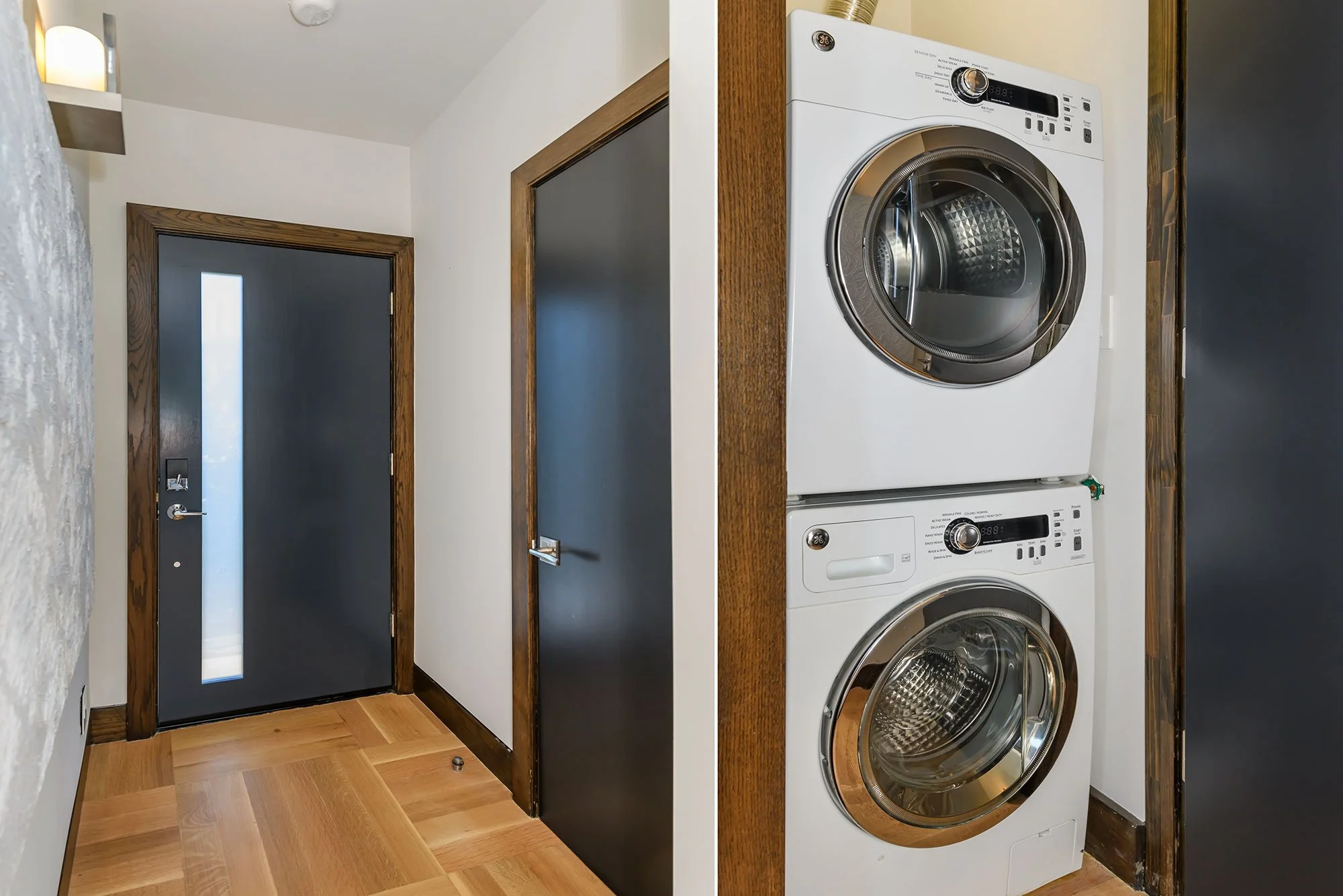 A laundry area with a stacked washer and dryer set against a wooden-paneled side, adjacent to a doorway with a dark door featuring a vertical frosted window. The room has wood flooring and white walls.
