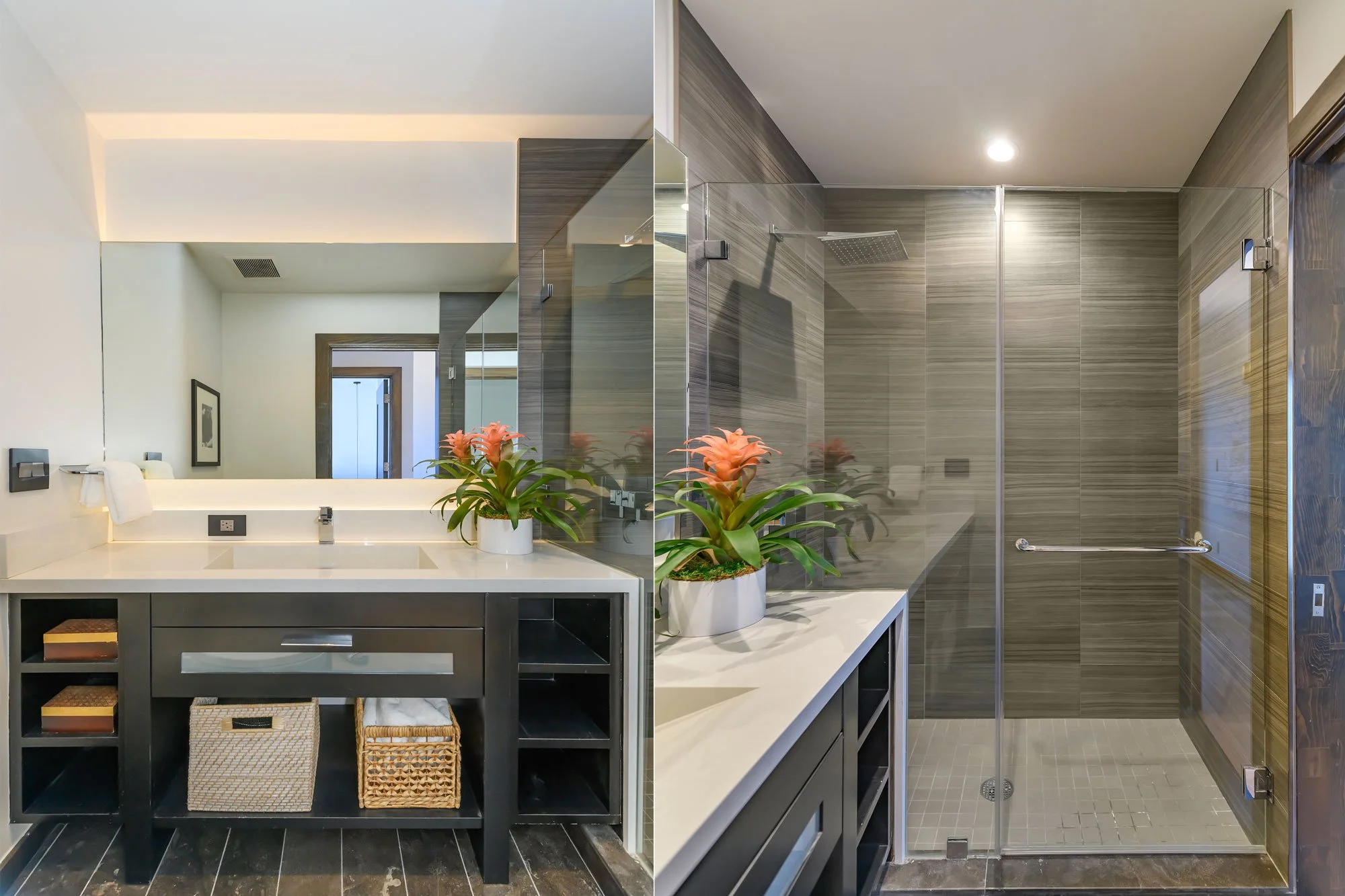 Modern bathroom with a large mirror, a vanity with a black finish, a white countertop, pink flowers in a white pot, and a walk-in shower with gray tiles and a glass door.