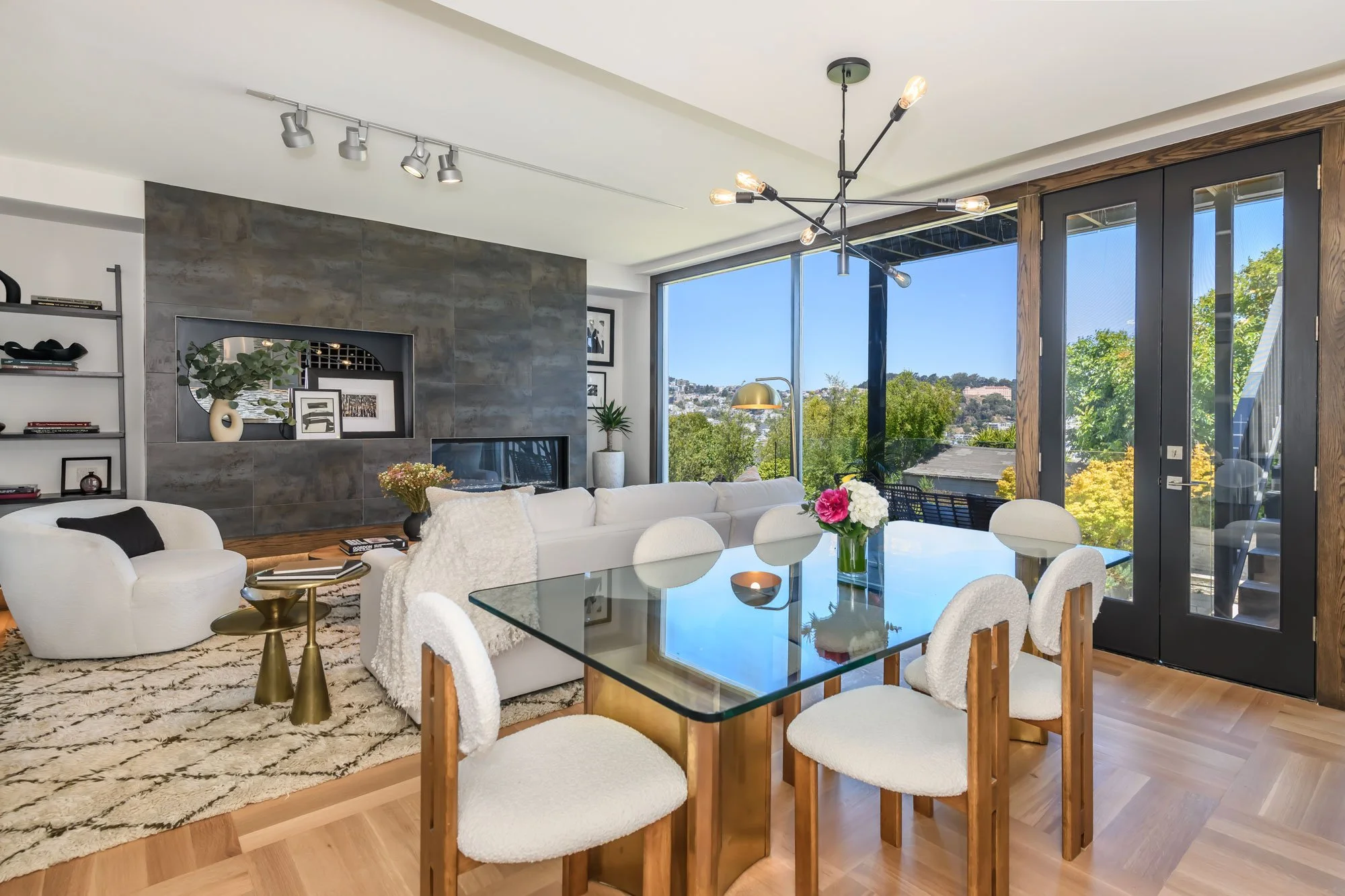 Modern living room with large windows showing trees and blue sky, white and beige furniture, a glass dining table with white chairs, a dark gray accent wall with decorative shelves and framed art, and a black and brass chandelier.
