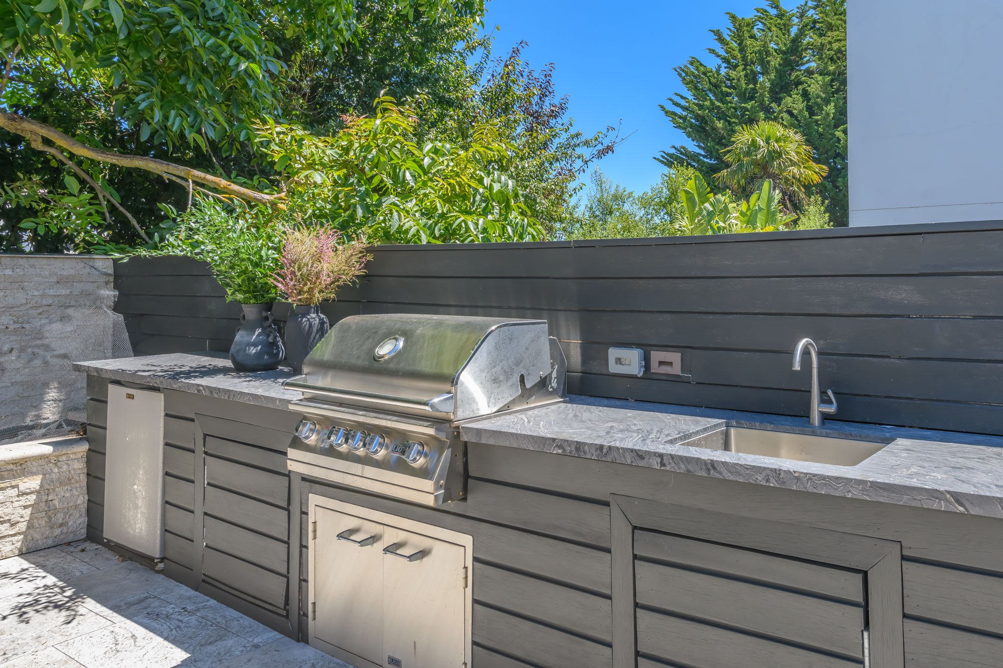 Outdoor kitchen with stainless steel barbecue grill, black potted plants, and a dark gray wooden privacy wall, surrounded by lush green trees and a clear blue sky.
