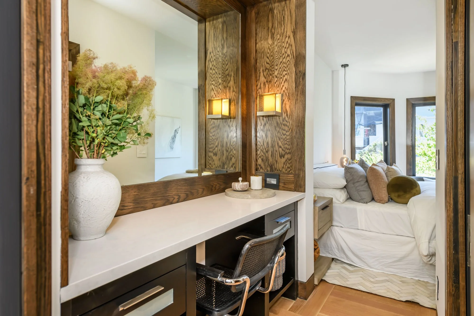 A bedroom with a wooden frame and a mirror on the wall, a white dresser with a vase of dried flowers, candles, and a small bowl. In the background, a bed with pillows and a bedside lamp, two windows showing trees outside.