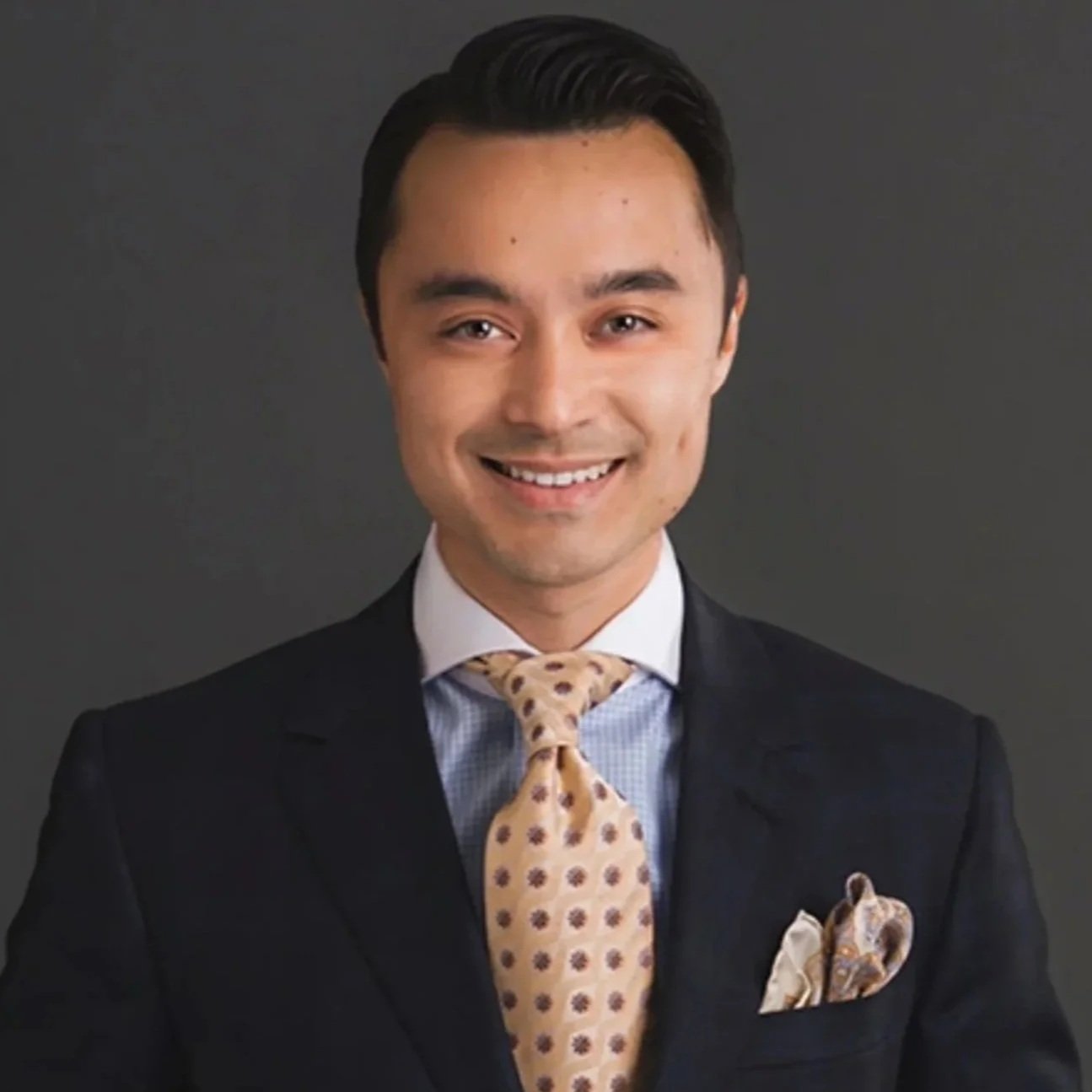 Portrait of a young man in a business suit with a patterned tie and pocket square, smiling, against a dark gray background.