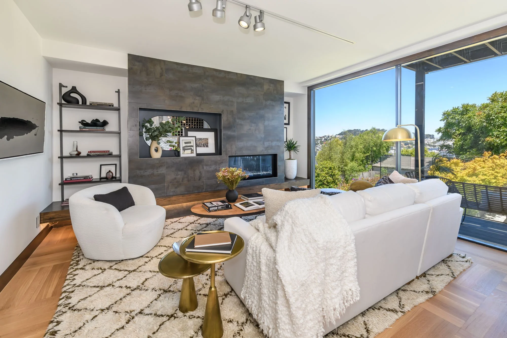 Modern living room with white sofa, black and white artwork, large window showing trees and cityscape, wooden flooring, and a fireplace with decorative shelves.