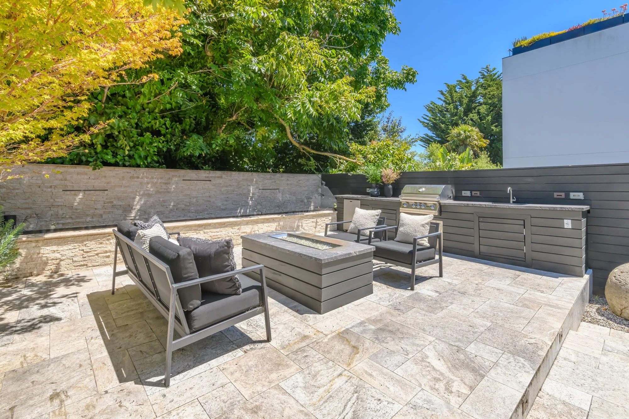 Outdoor patio with seating area, barbecues, and lush green trees under a clear blue sky.